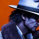 A man wears a white hat and suit in front of an orange background.